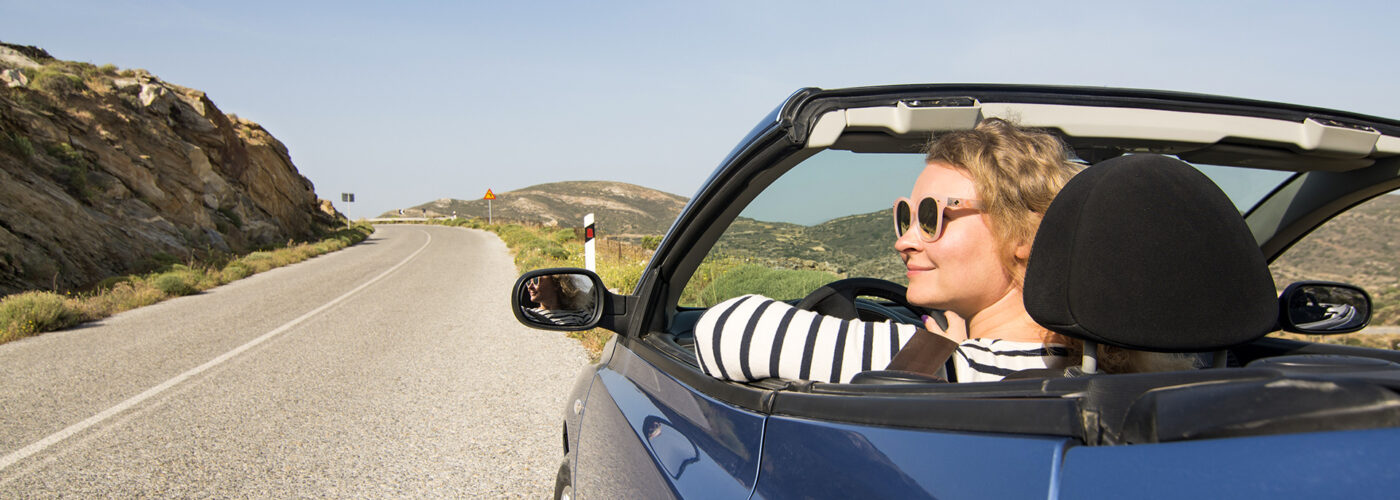 Young blonde woman driving in convertible blue car without roof on mountain road in Naxos island, Greece.