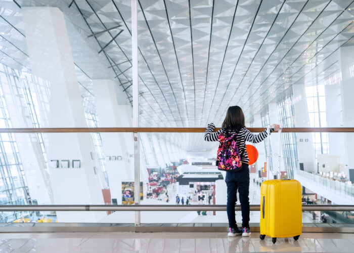Child overlooking an airport terminal from above next to a yellow rolling suitcase