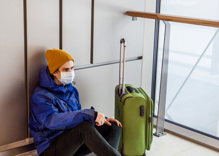 boy with a green suitcase is sitting on floor in waiting area wearing protection mask