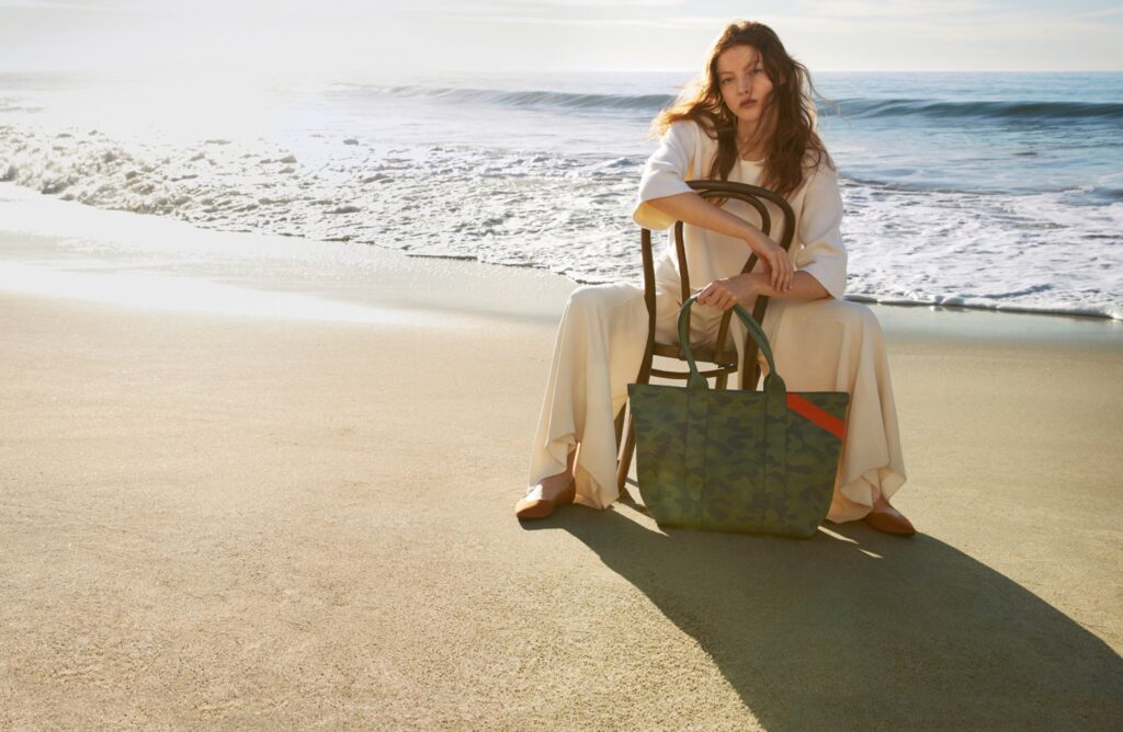 Woman sitting backwards on a chair on the beach holding a bag from the Rothy's bag collection