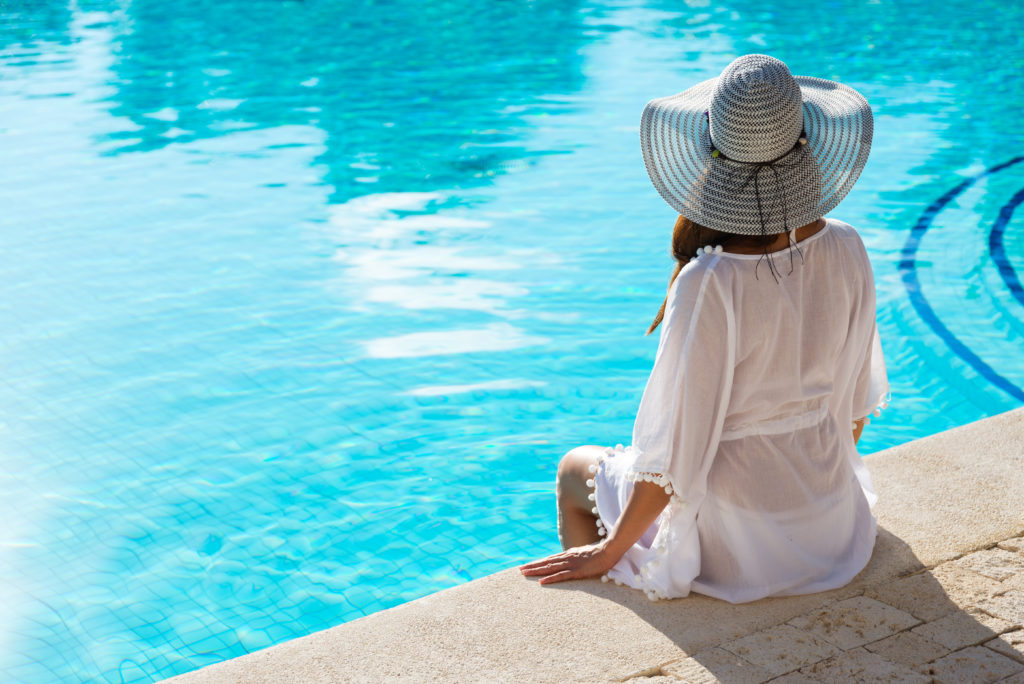 A woman facing away from the camera in a white swimsuit cover up and hat looking out over a resort pool