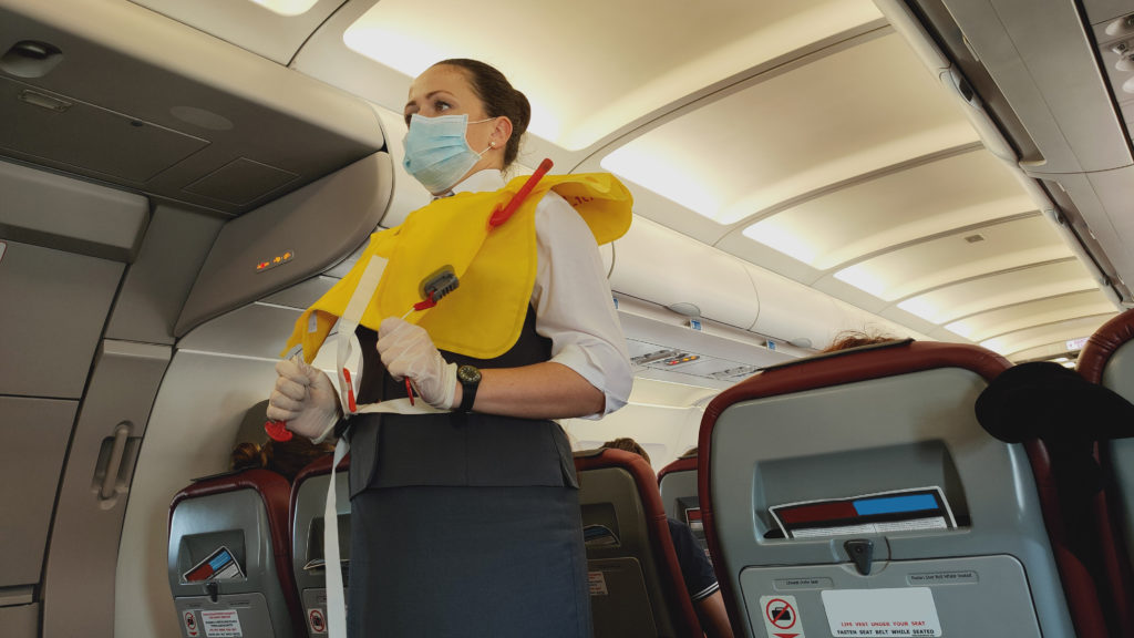 Masked flight attendant demonstrates how to inflate a life vest