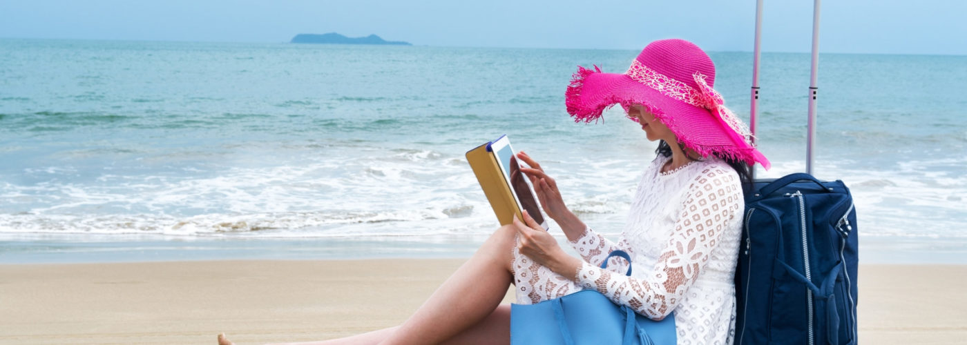 Tourist reclining against her luggage on the beach, reading on her iPad