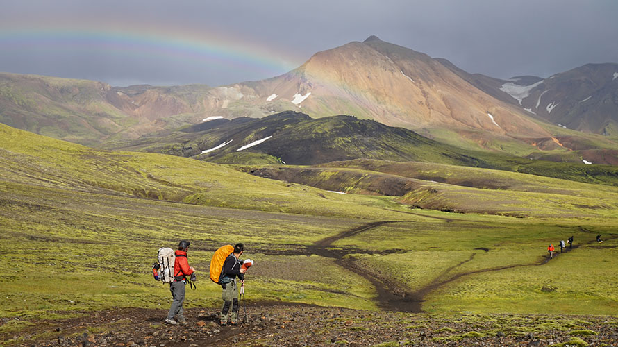 Six hikers, two in foreground and three further away, on a hiking path through rolling hills with a rainbow overhead