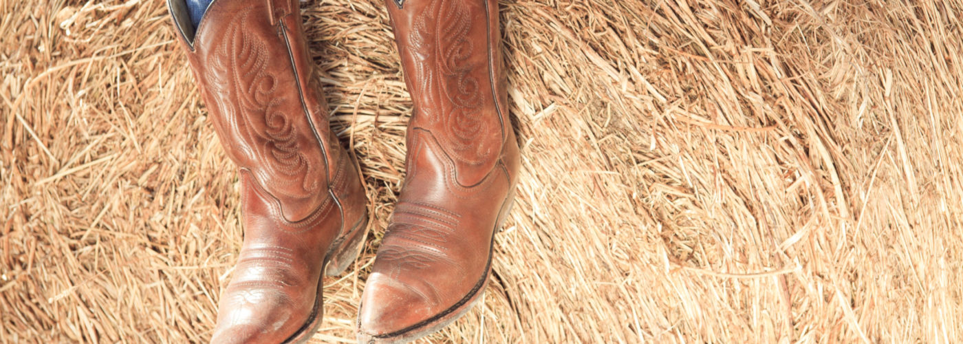 Person from the knees down wearing cowboy boots resting on a pile of hay