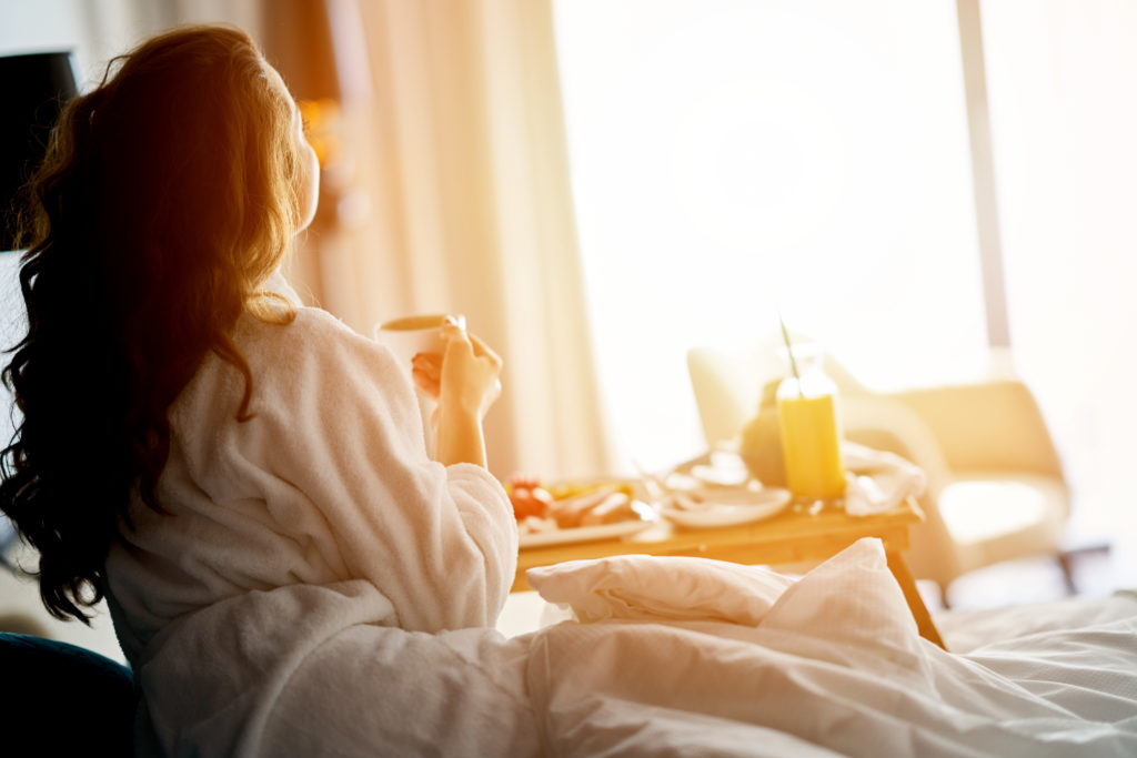Woman looking out a bright window, having breakfast in bed at hotel