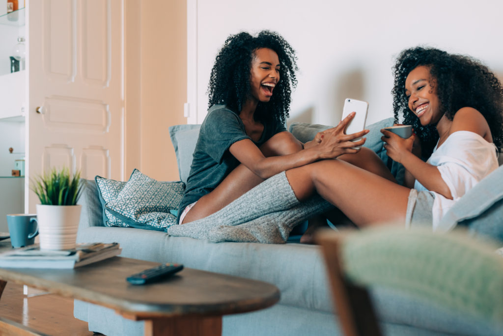 Two women laughing at phone screen and drinking coffee on couch