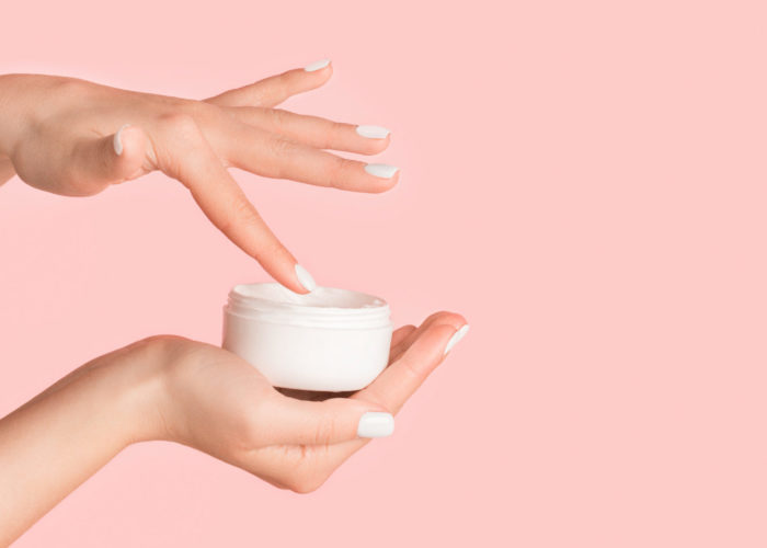Close-up of hands dipping fingers into a small jar of moisturizer on a light pink background
