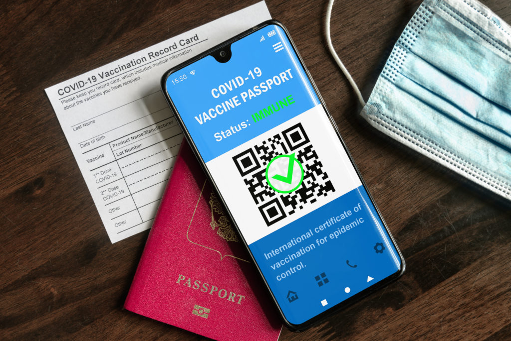 COVID-19 vaccination card, vaccine passport on phone, passport, and face mask on wooden table
