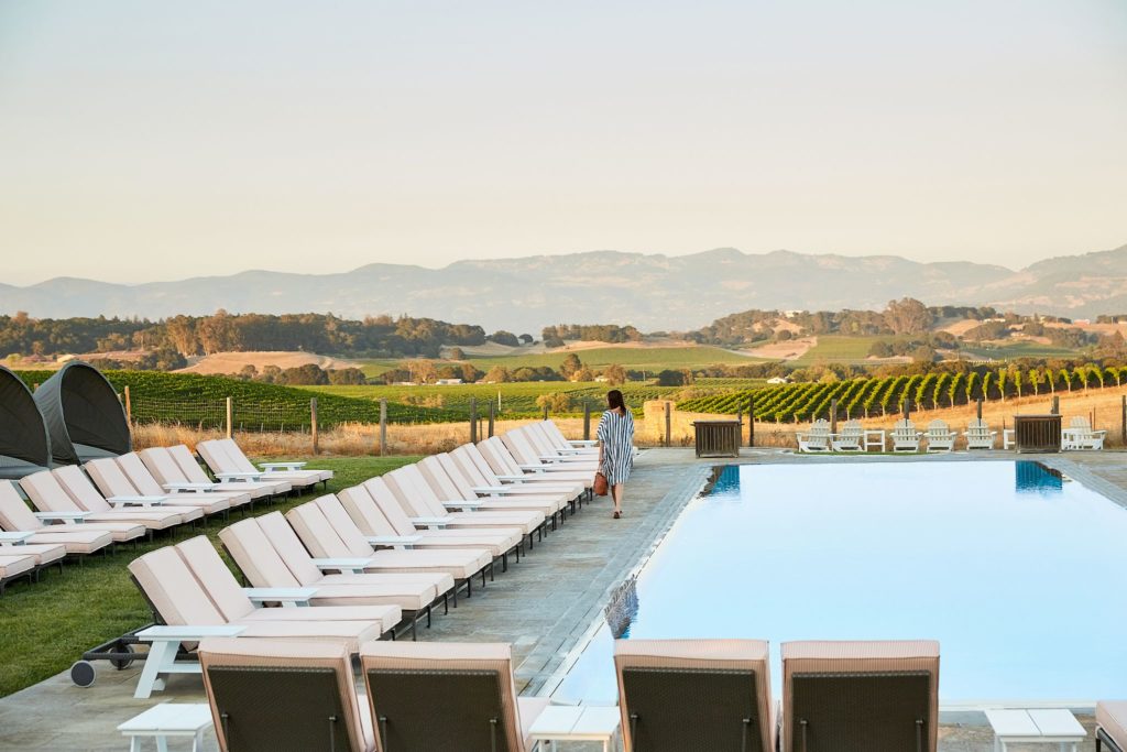 Woman walking on a pool deck next to a pool and a line of pool chairs with a vineyard in the background at the Carneros Resort and Spa