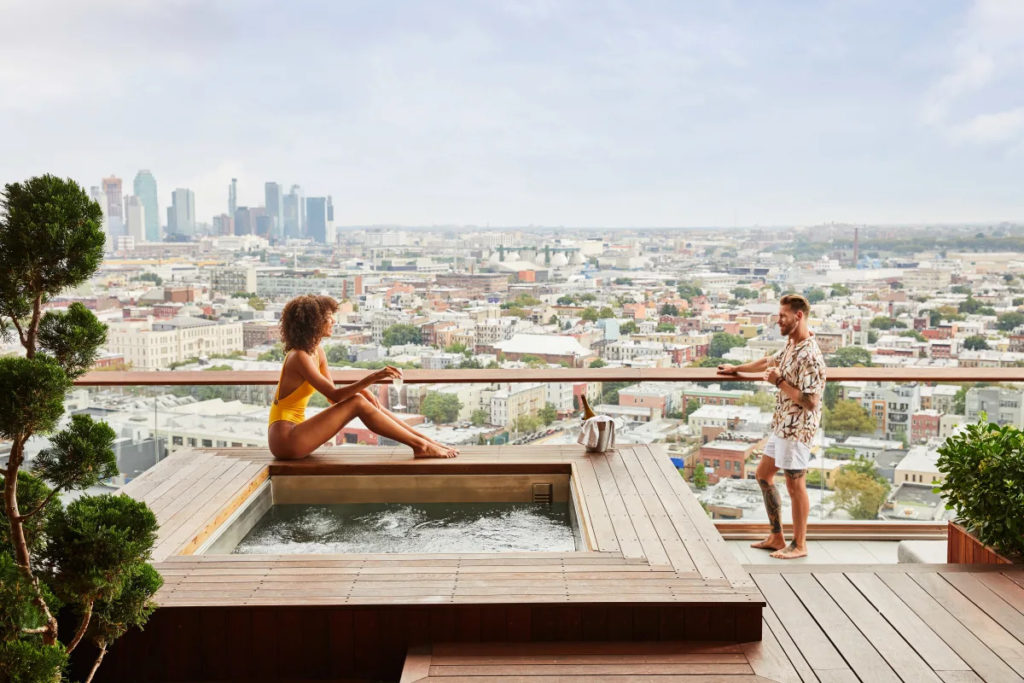 Two people relaxing around the wooden hot tub on a deck overlooking a cityscape at The William Vale, New York 