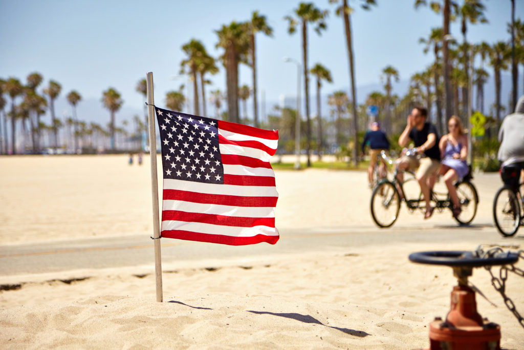 People riding bikes on Venice Beach in the background with an American Flag in the sand in the foreground