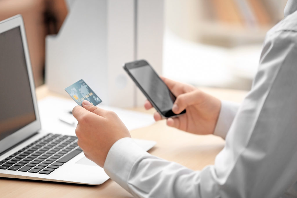 Close up of person holding a credit card in one hand and a smartphone in the other hand while working at a laptop
