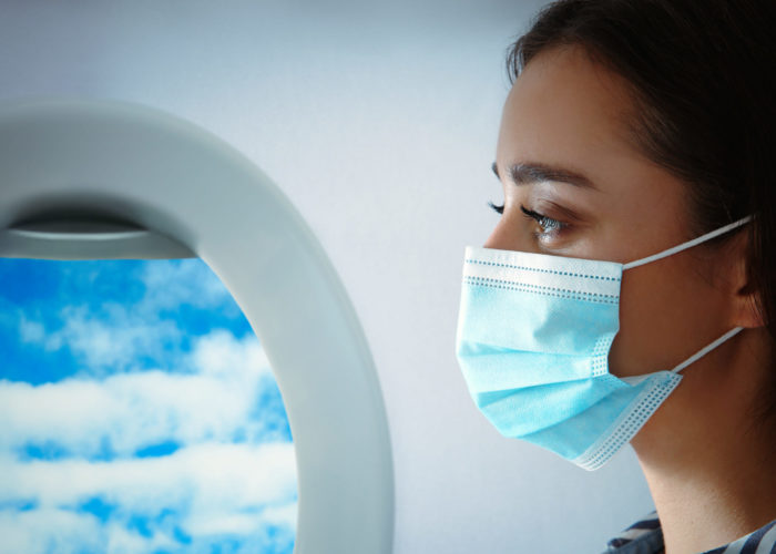 Close up of woman wearing a medical face mask on a plane next to an open window showing a blue sky with white clouds