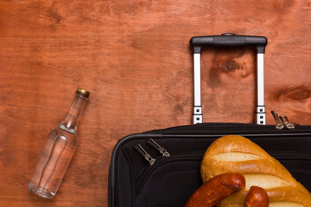 Top down view of a suitcase with bread and sausage on top, next to a glass bottle of clear liquid