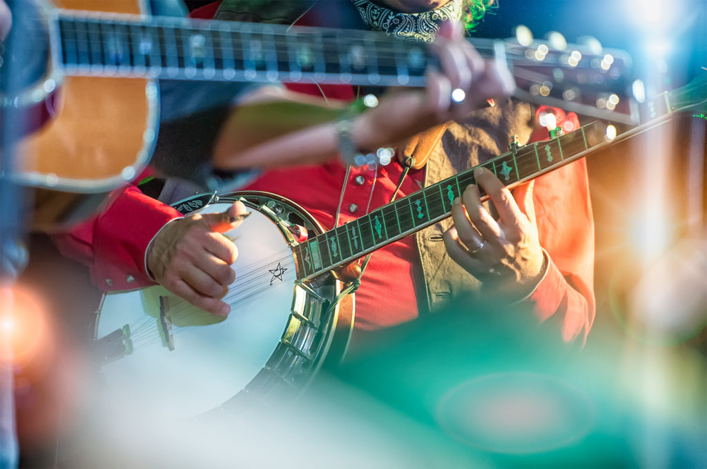 Close up of the hands of a banjo player strumming a banjo with a bandmate playing guitar in the foreground, out of focus