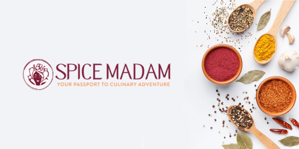Several spices in wooden bowls and spoons on a grey background next to the Spice Madam logo