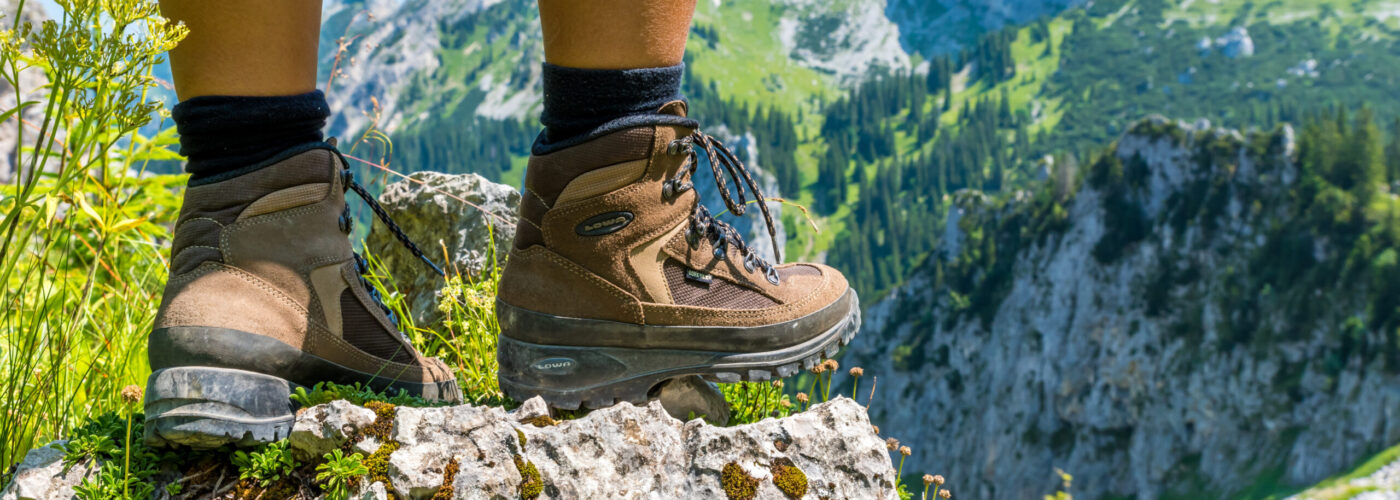 Close up of person from the knees down, wearing hiking boots and standing on a cliff overlooking a green mountainous landscape