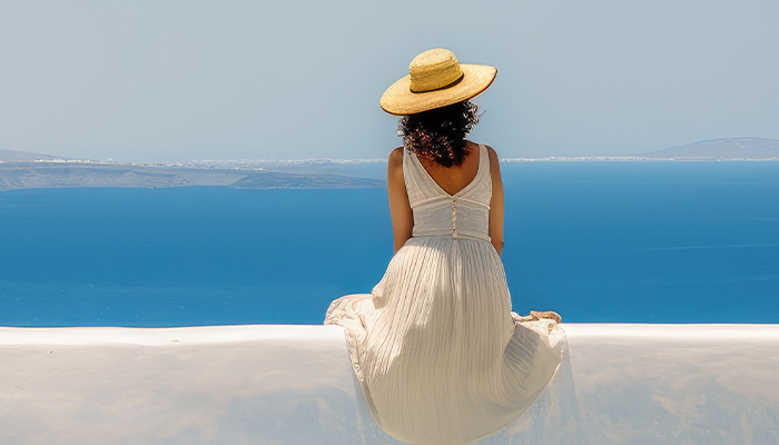 Beautiful young woman sitting on wall looking at stunning view of Mediterranean sea and Santorini village, Greece, Europe. Lifestyle woman with straw hat wearing green dress enjoy landscape view.
