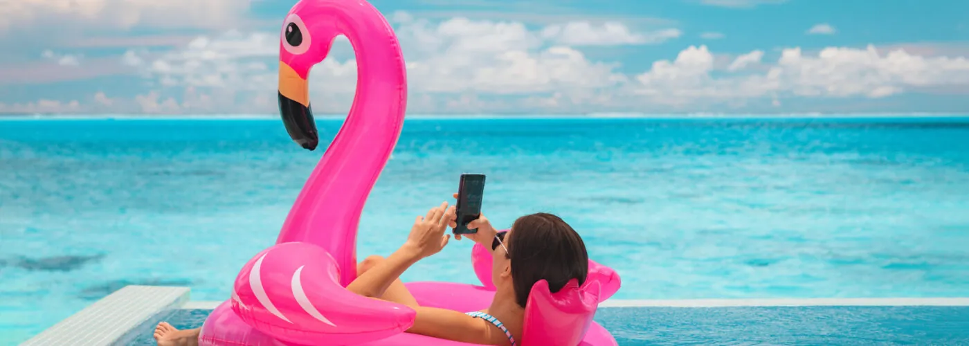 Woman lounging on inflatable flamingo in pool overlooking the ocean scrolling on smartphone